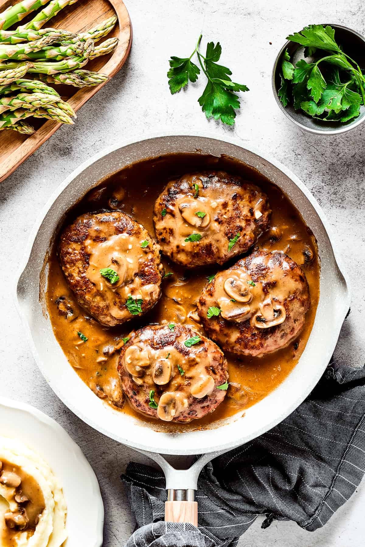 Four salisbury steaks in gravy, in a skillet. Vegetables, herbs, and a cloth napkin are arranged around the skillet.