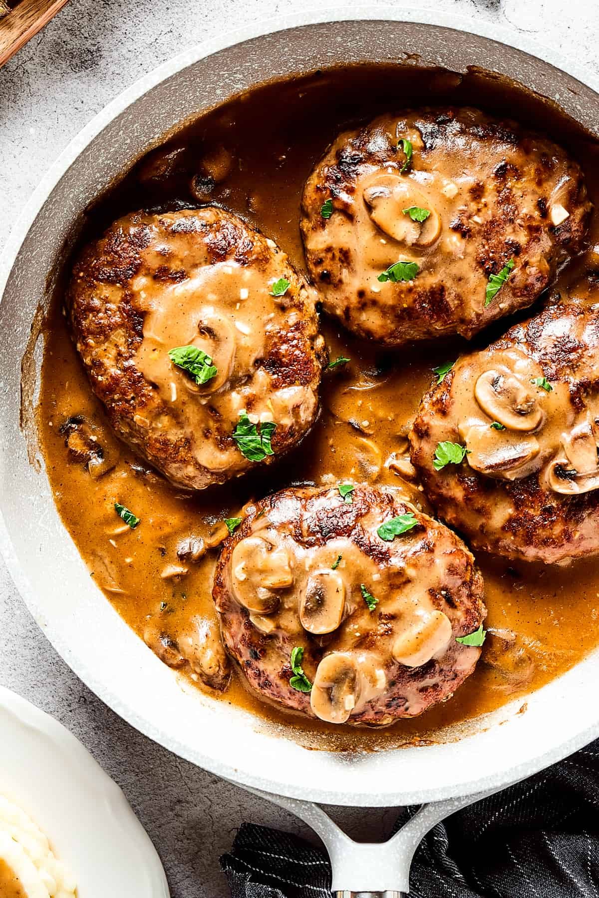 Four salisbury steaks in gravy, in a skillet. Vegetables, herbs, and a cloth napkin are arranged around the skillet.