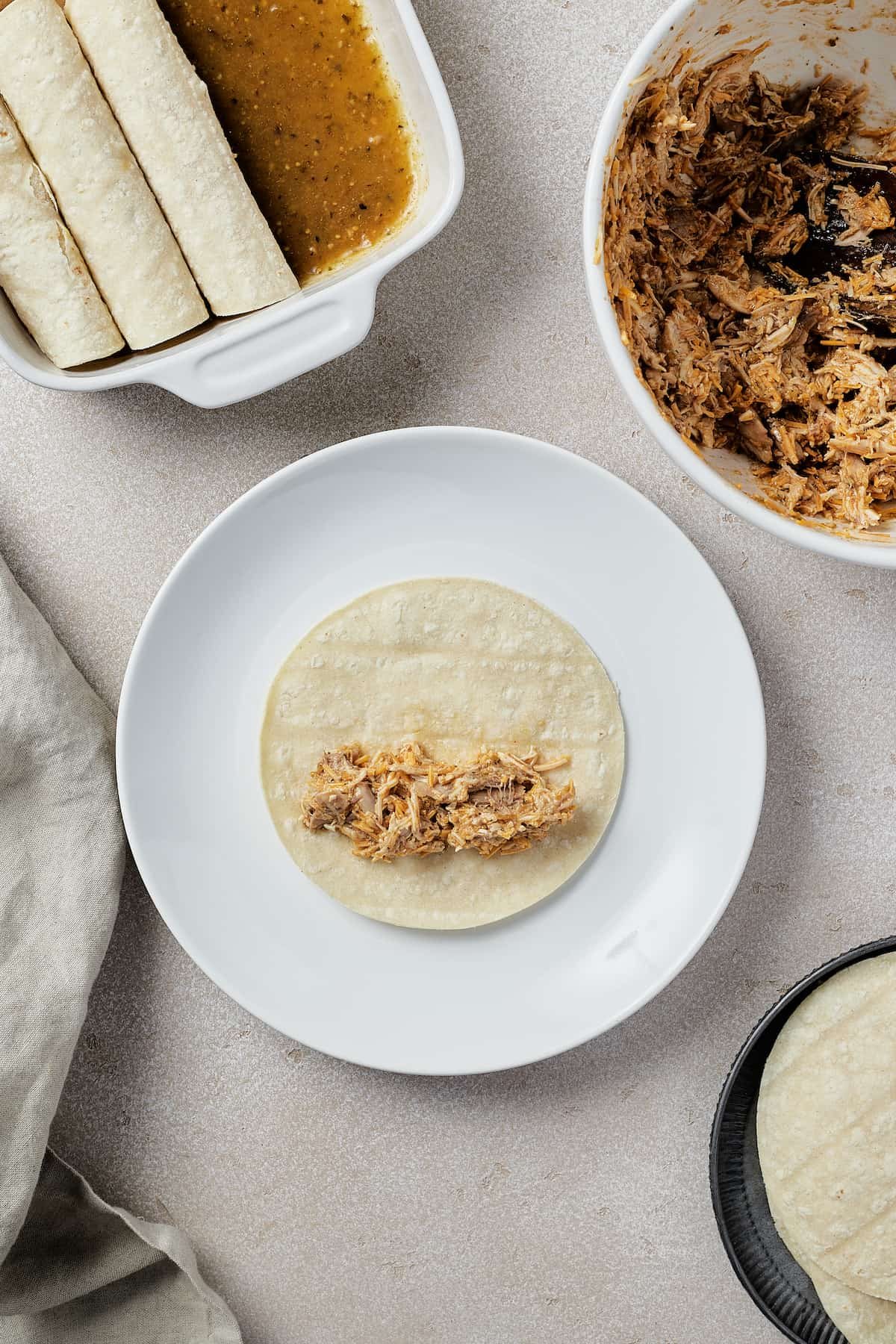 A small corn tortilla on a plate with a spoonful of chicken filling arranged on it. A dish of enchiladas, a bowl of filling, and more tortillas are visible in the edges of the frame.