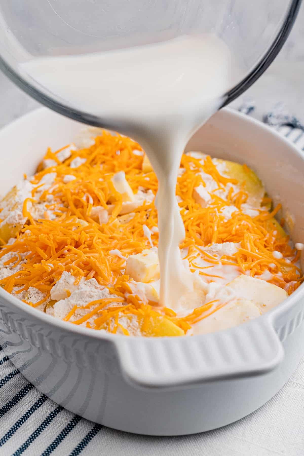 Pouring milk over sliced potatoes and shredded cheese in a baking dish.