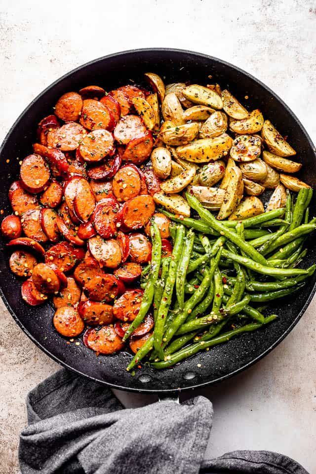 grey linen kitchen towel next to a black skillet filled with andouille sausage slices, potatoes, and green beans
