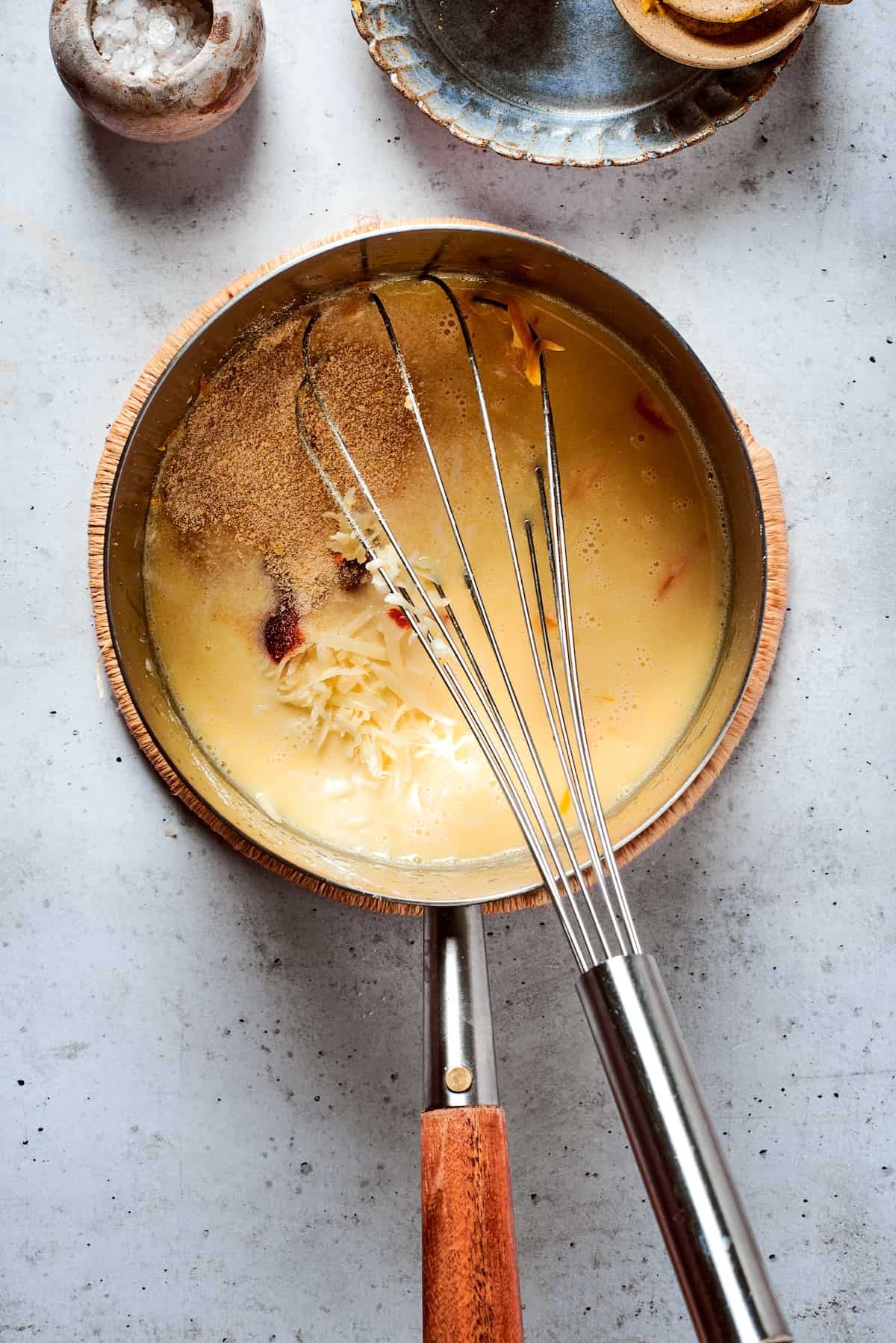 A whisk whisks a cheese sauce in a metal pot.