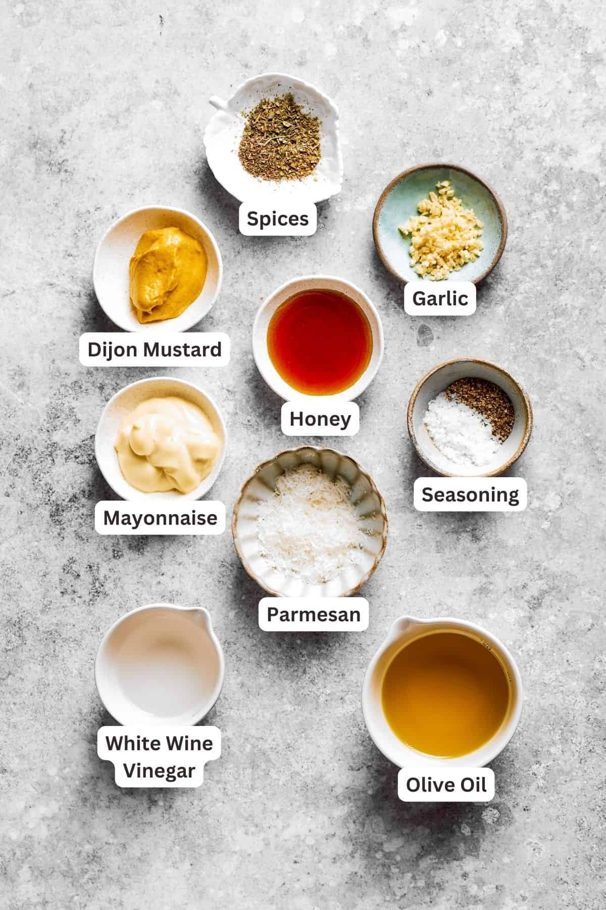 The ingredients for the dressing for spaghetti salad are labeled and portioned: dijon mustard, vinegar, honey, dried herbs, parmesan, mayonnaise, garlic, seasoning salt.