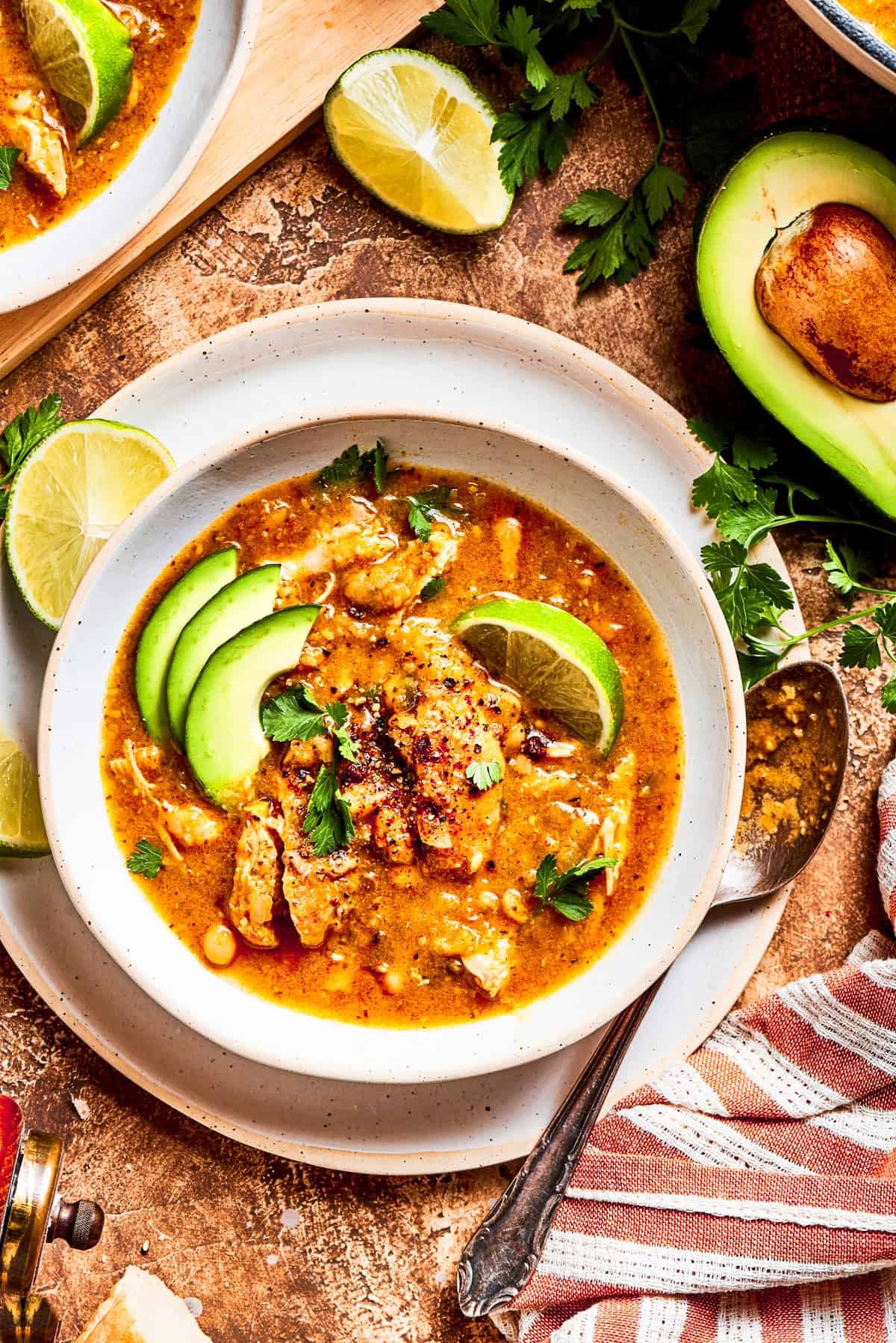 Spicy chicken soup in a bowl garnished with avocado slices and lemon wedges.
