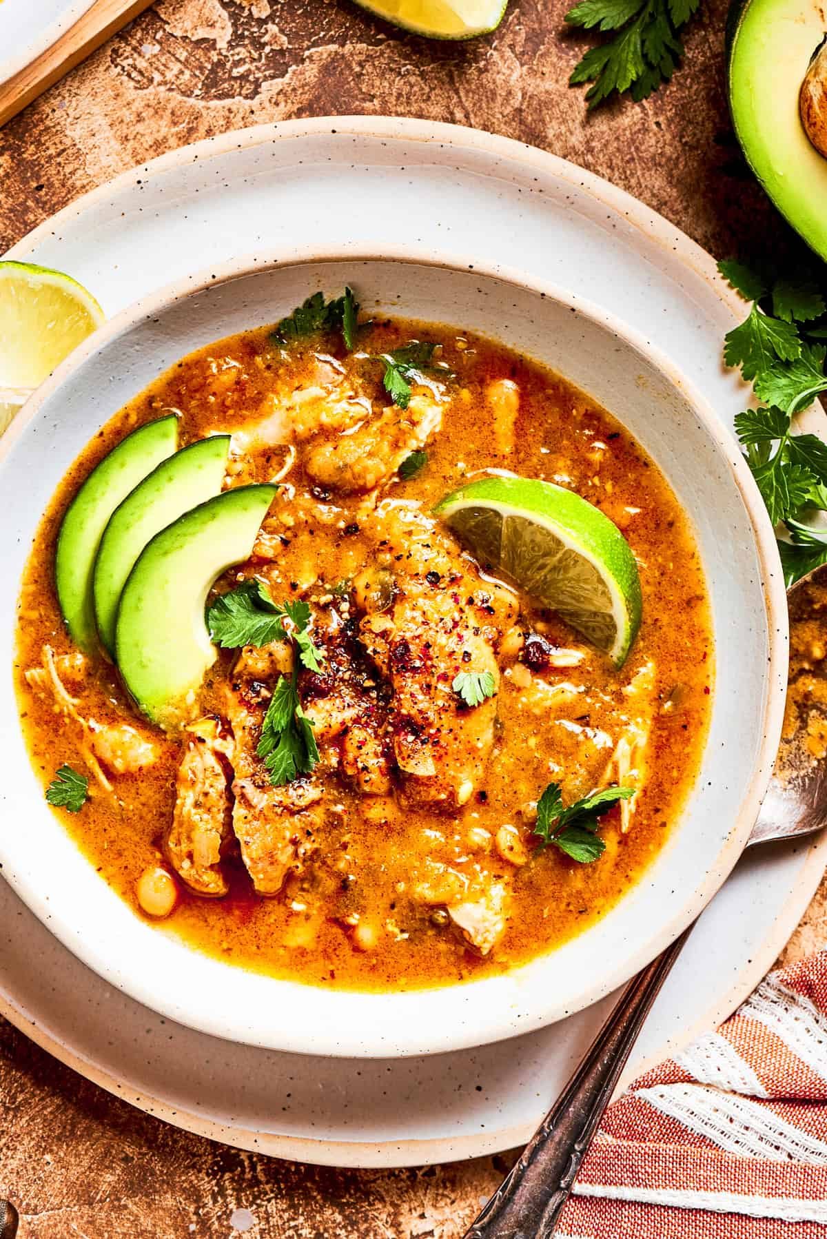 Spicy chicken soup in a bowl garnished with avocado slices and lemon wedges.
