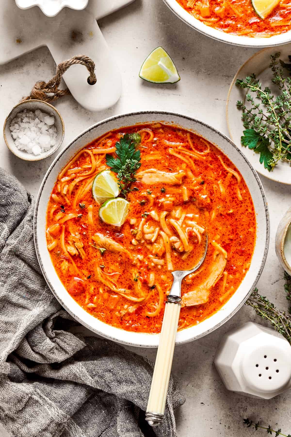 Two bowls of spicy chicken soup are shown with toppings arranged around them.