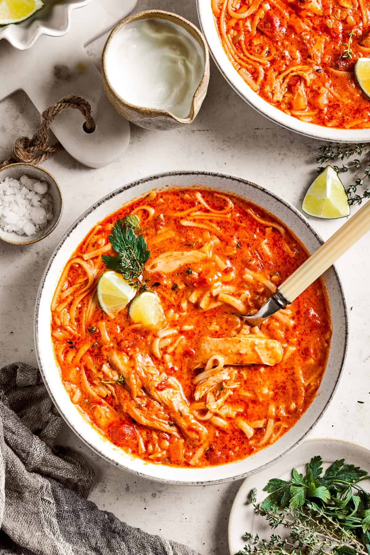 Two bowls of spicy chicken soup are shown with toppings arranged around them.