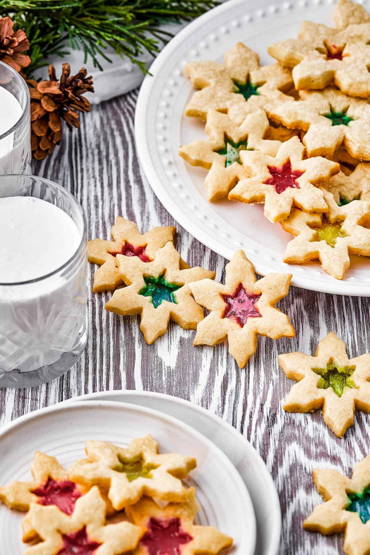 Two plates of stained glass cookies, with extra cookies arranged artistically on the table, along with a glass of milk.