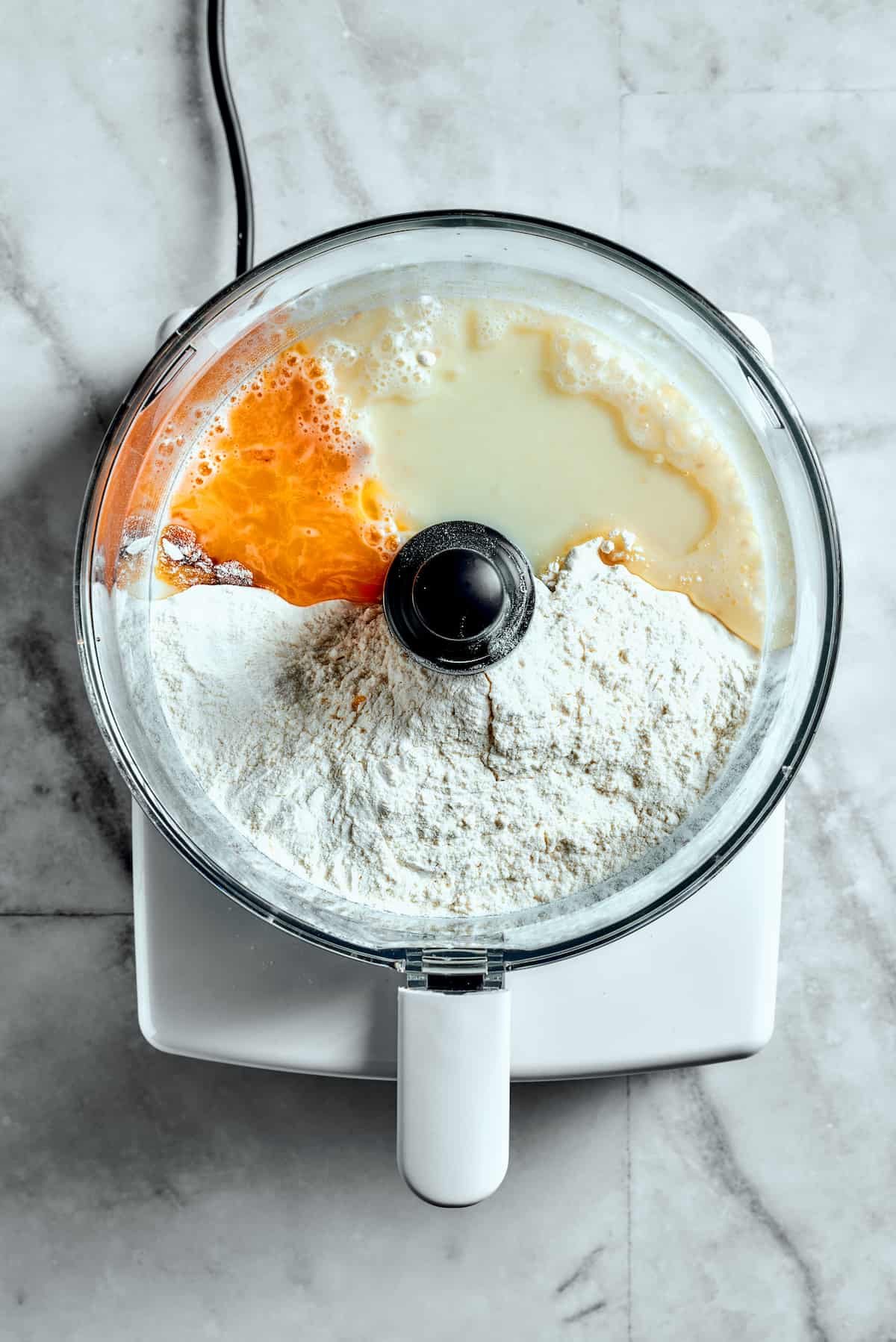 Overhead view of flour, egg yolk, milk, and yeast in a food processor.