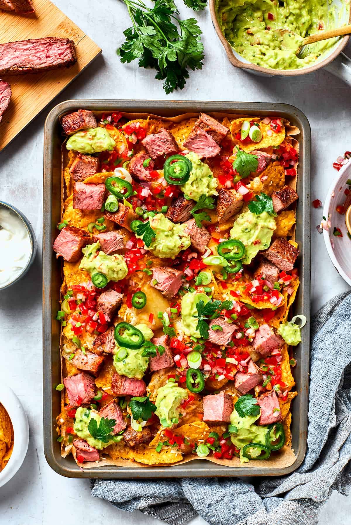 A metal baking sheet of steak nachos is surrounded by ore steak and toppings.