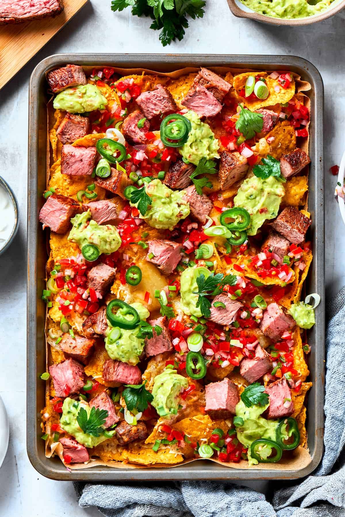 A baked pan of steak nachos is topped with dollops of guacamole and chunks of steak.