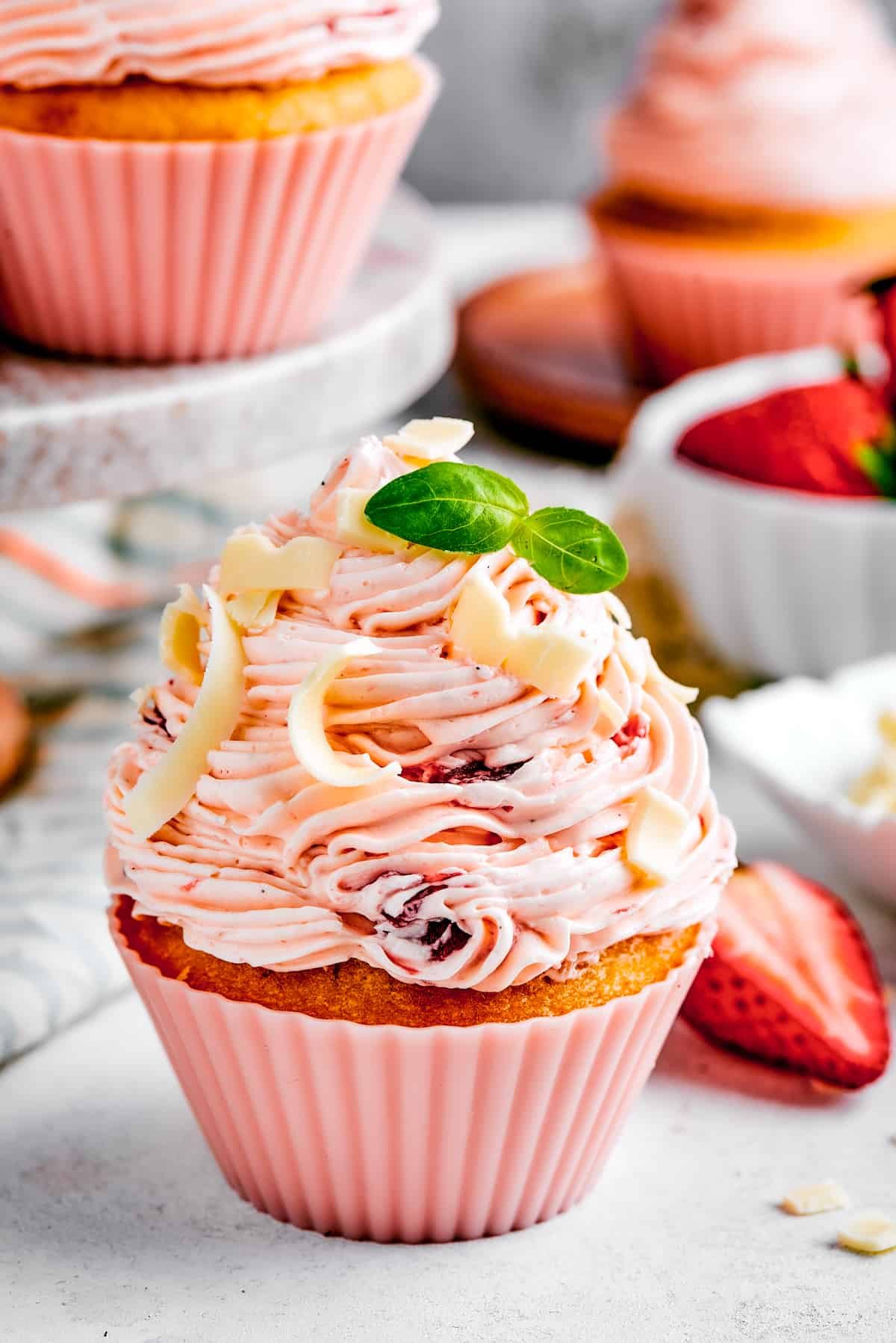 A strawberry cupcake in a pink cupcake liner with a tiny sprig of mint on top. More cupcakes are visible in the background of the shot.