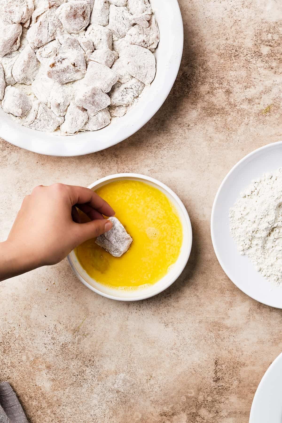 A chicken breading station with breaded chicken, egg wash, flour, and pieces of floured chicken on a plate.