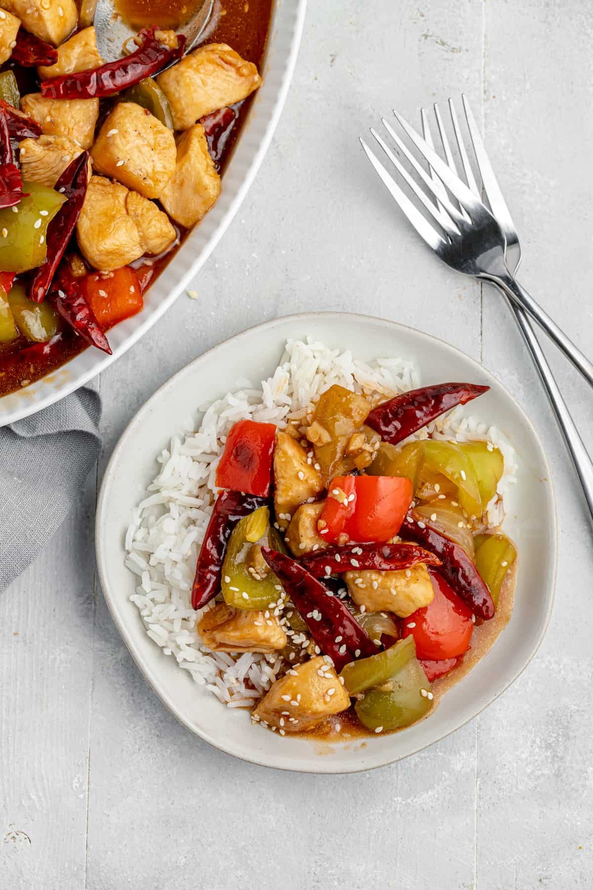 A serving of Szechuan chicken stir-fry over rice with a serving platter and two forks nearby on the table.