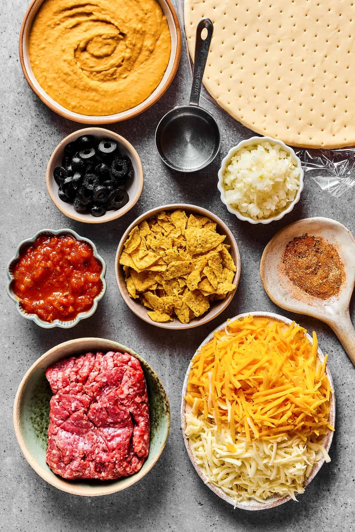 From top: refried beans, pizza crust, black olives, water, onion, salsa, crushed tortilla chips, taco seasoning, ground beef, cheese.