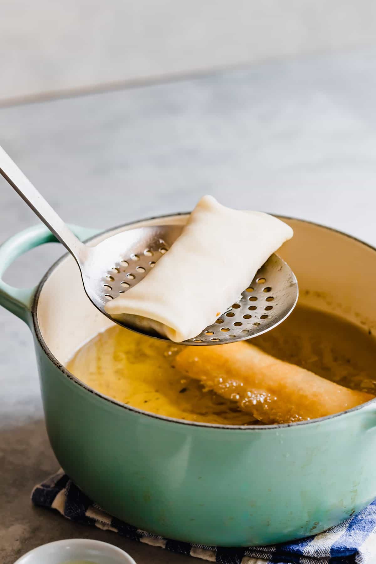 A Slotted Spoon Holding an Uncooked Egg Roll Over a Teal Frying Pan