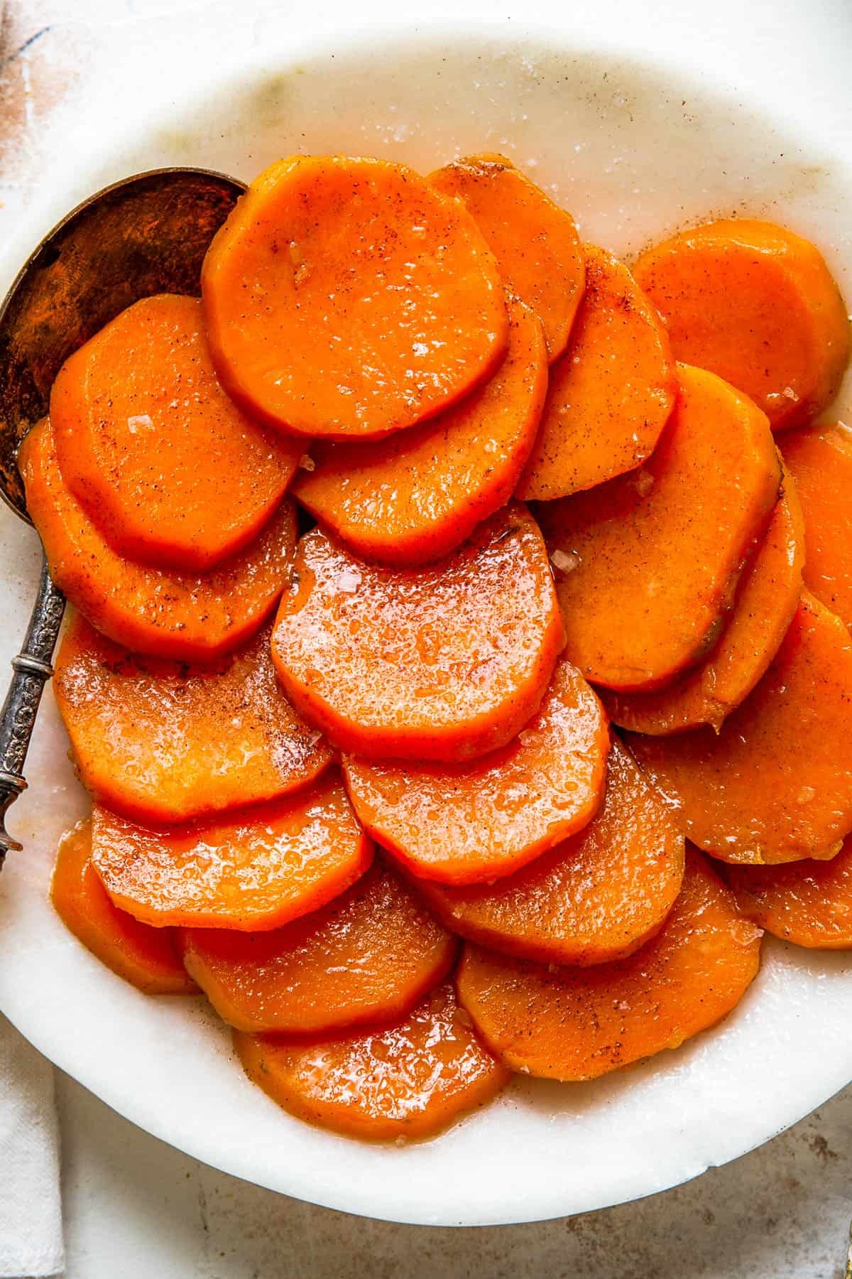 A white serving dish with sliced candied sweet potatoes and a serving spoon.