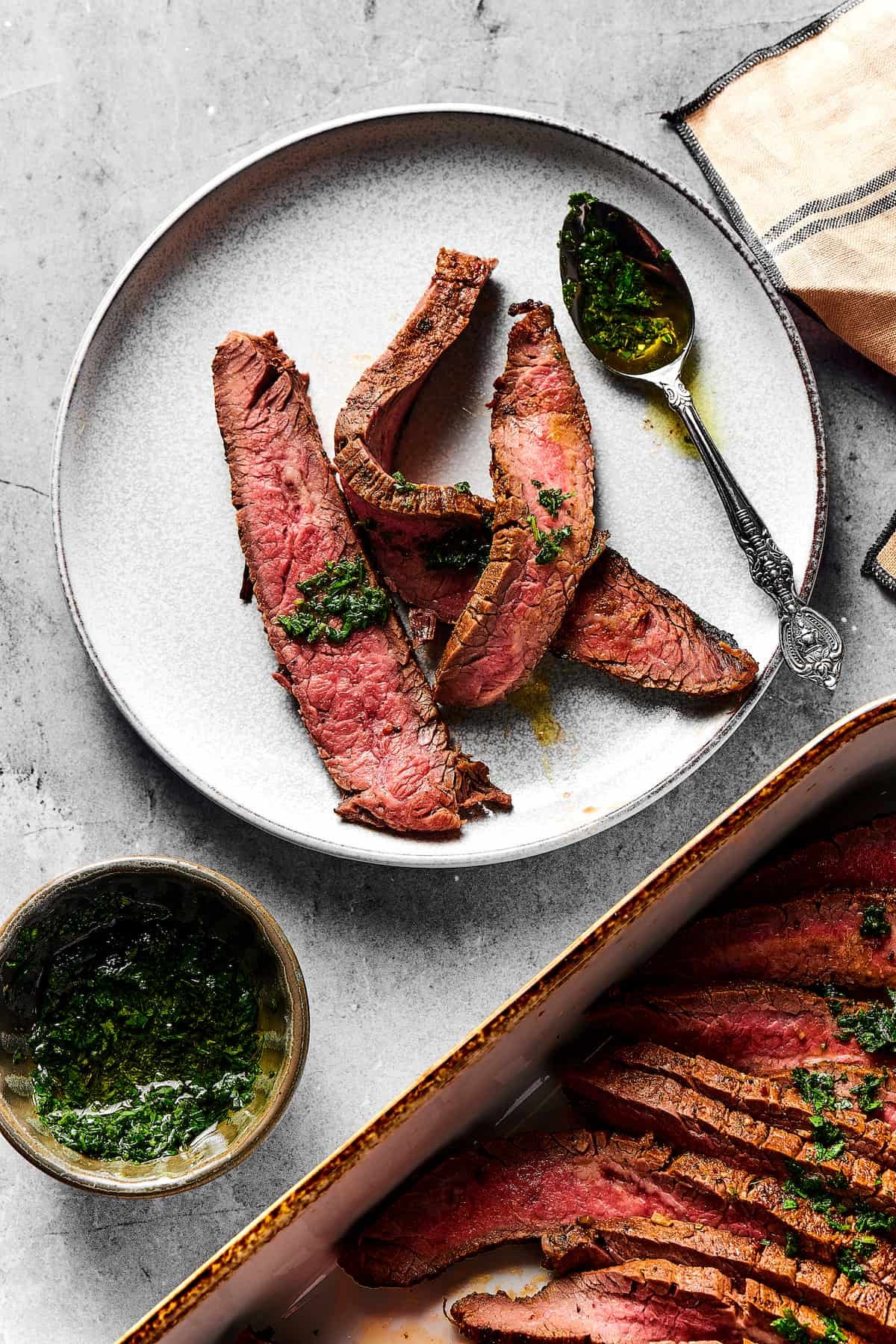 A small plate with three strips of medium-rare grilled carne asada on it. Nearby is a large platter of the steak slices, a cup of sauce, and a spoon.