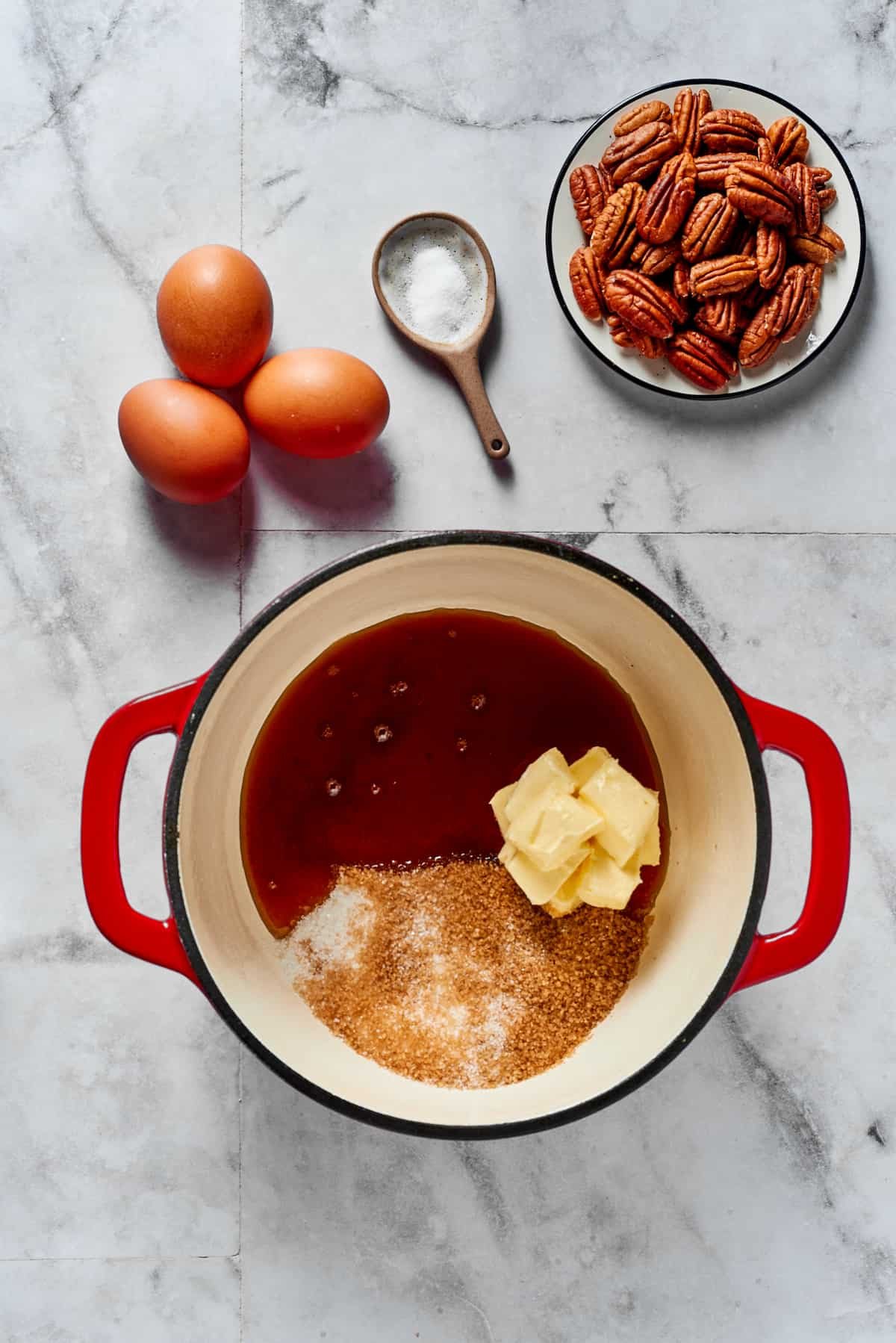 A heavy pot with butter, sugar, and syrup inside. Eggs, salt, an pecans are arranged in dishes on the work surface.