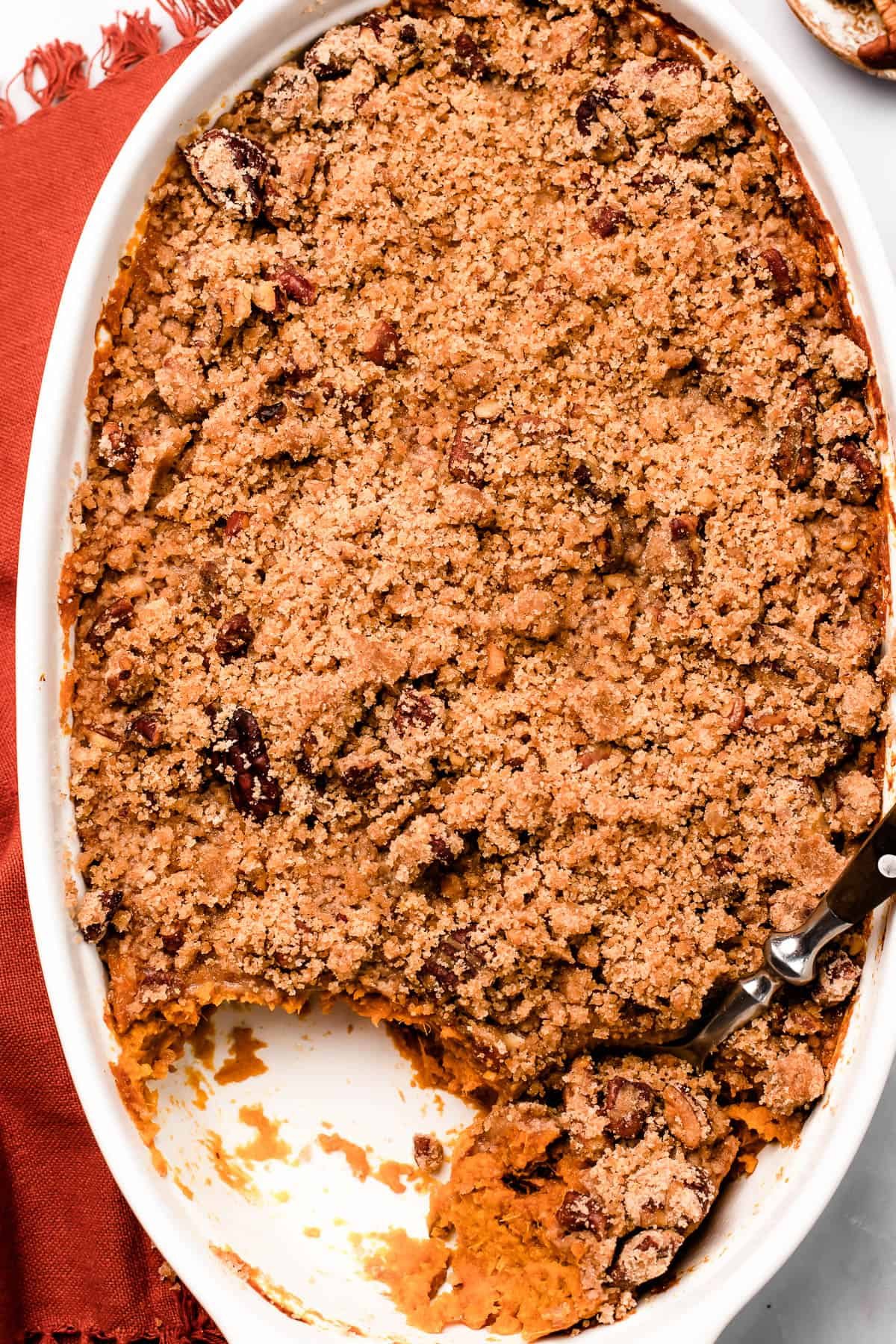 A baking dish of sweet potatoes and crumb topping, on a marble background. A serving of the casserole has been scooped out, and a spoon is resting in the dish.