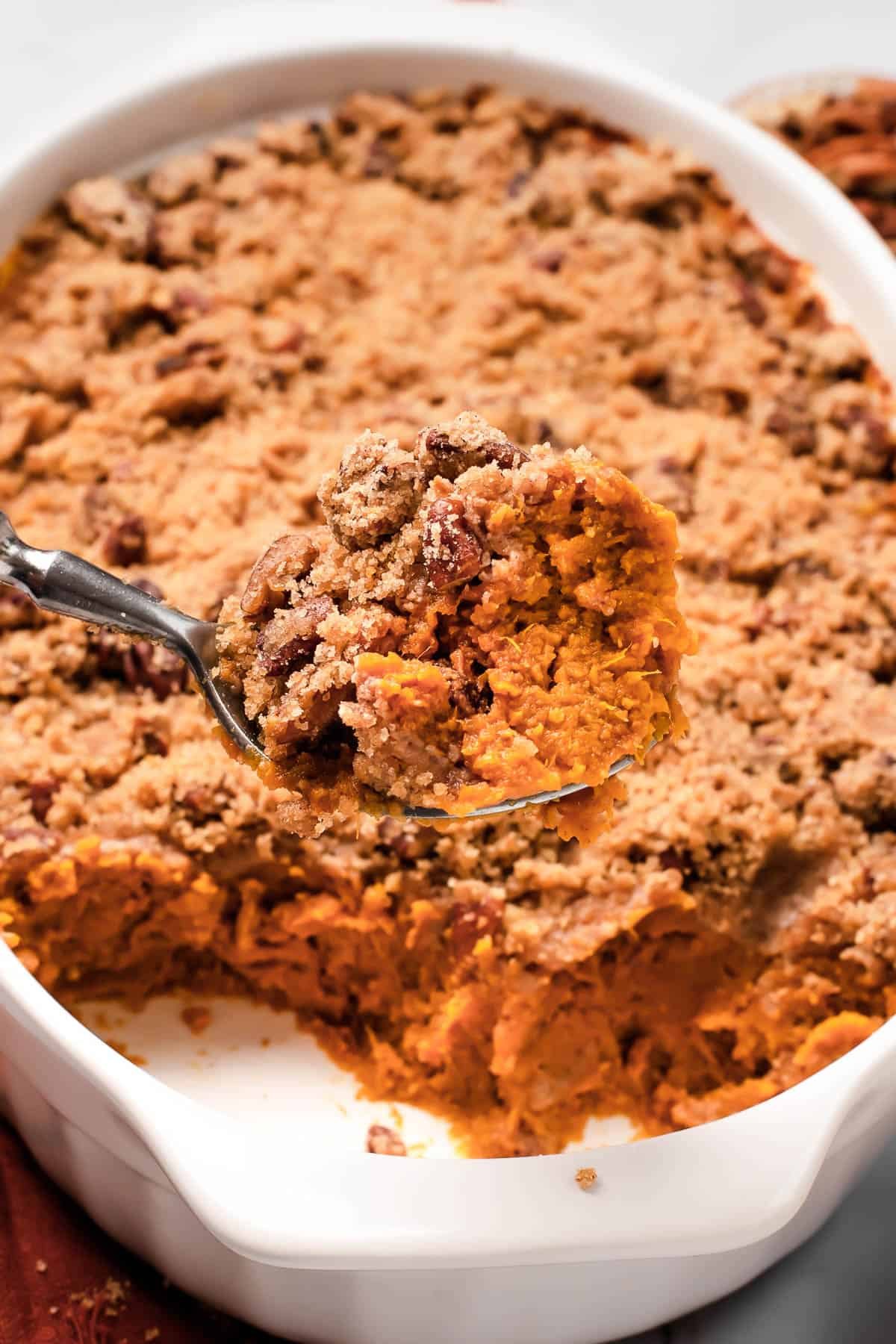A shot of the sweet potato casserole with a serving spoon lifting a portion out of the dish, showing the texture of the baked casserole mixture and crumb topping.