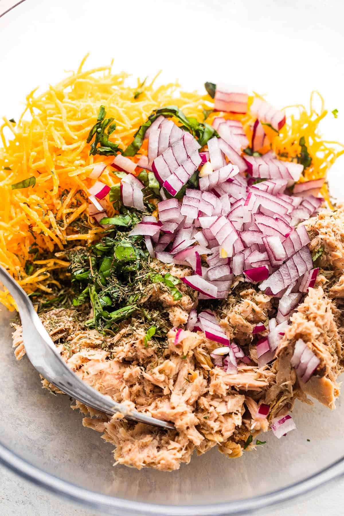 flaked tuna in a glass bowl with shredded cheddar cheese, diced red onions, and fresh herbs.