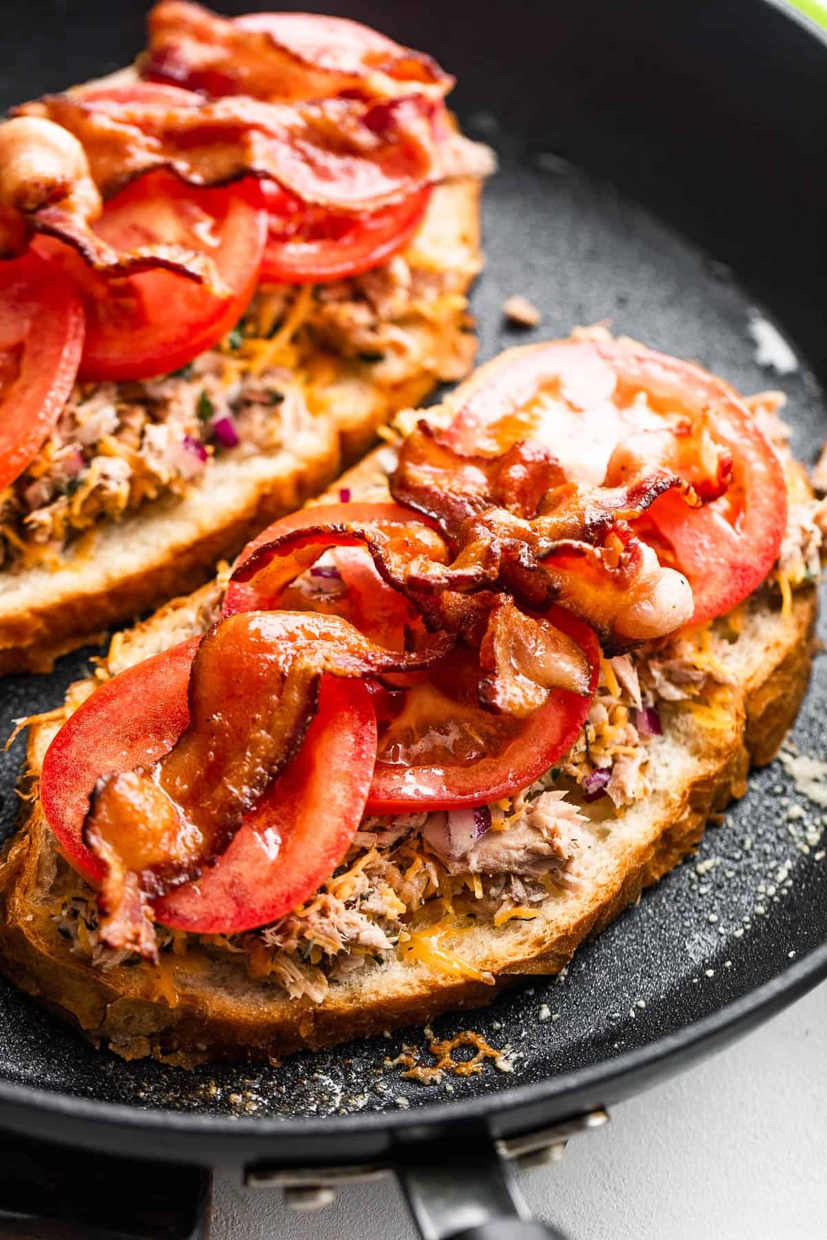 bread slices topped with flaked tuna, tomatoes, and bacon, all cooking in a black skillet.