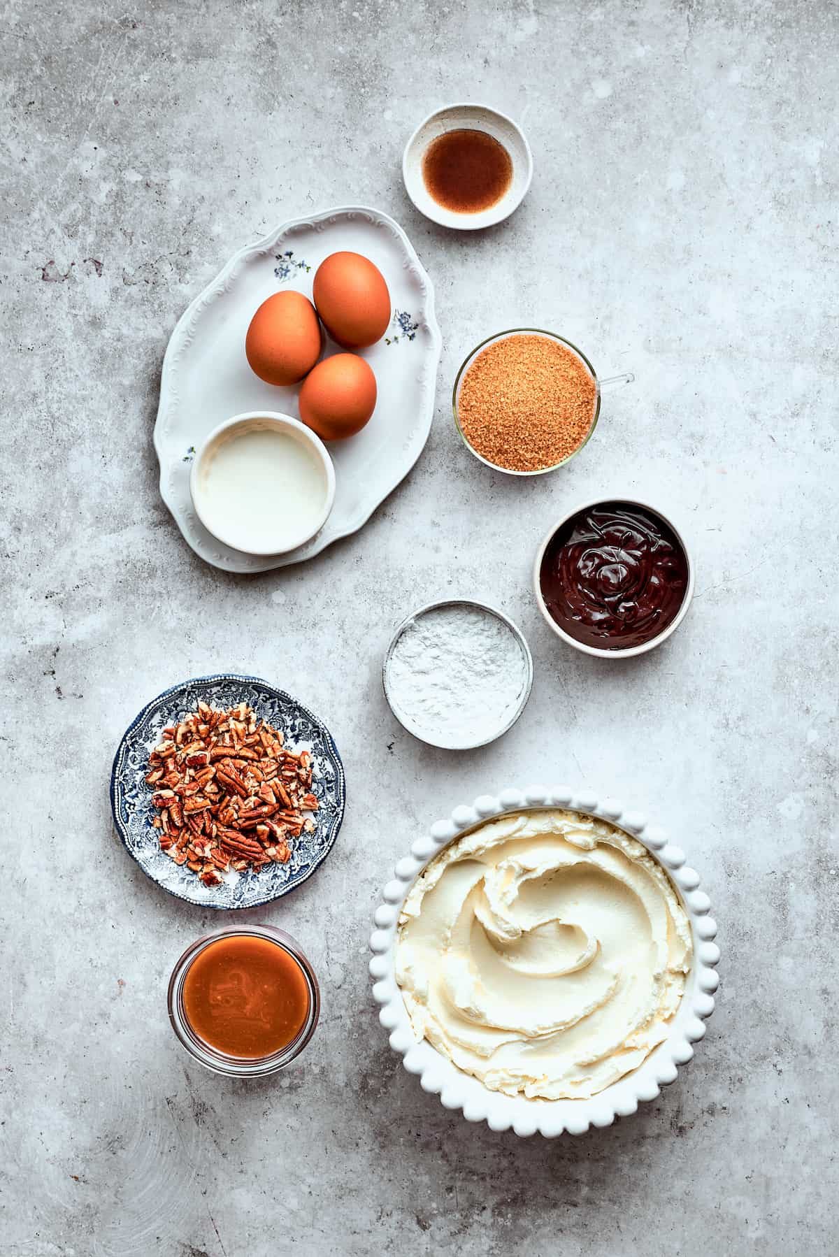 The ingredients for turtle cheesecake are shown portioned out on a white background: eggs. pecans, caramel, cream cheese, chocolate chips, brown sugar, vanilla, sour cream.