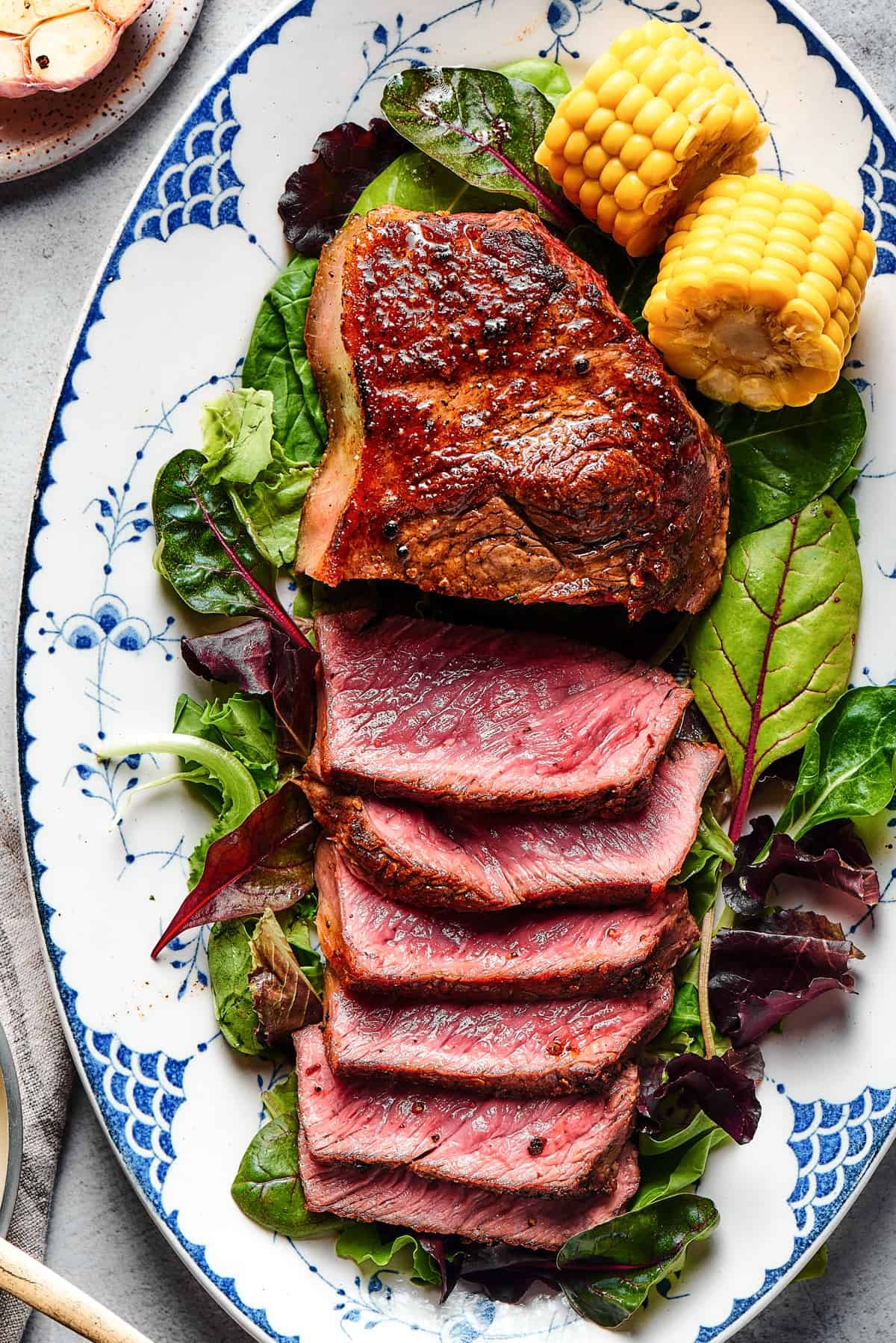A steak on a platter of greens, garnished with corn on the cob. About half the steak has been sliced and fanned out to show the rare interior.