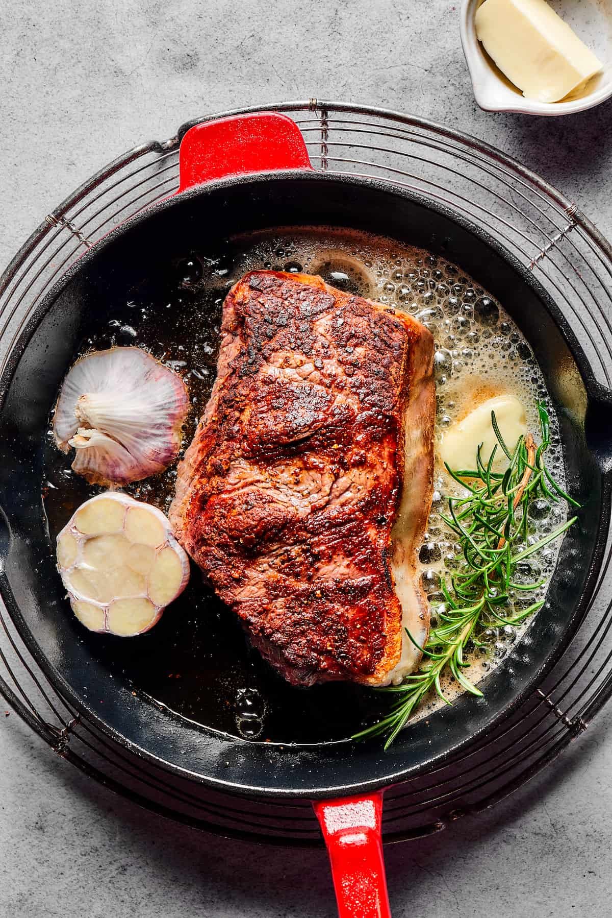 Sirloin steak in a cast-iron pan with melting butter, a sprig of rosemary, and a cut head of garlic.