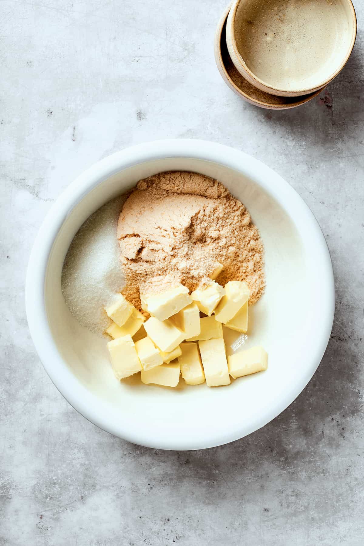 Butter, sugar, and brown sugar are added to a white bowl.