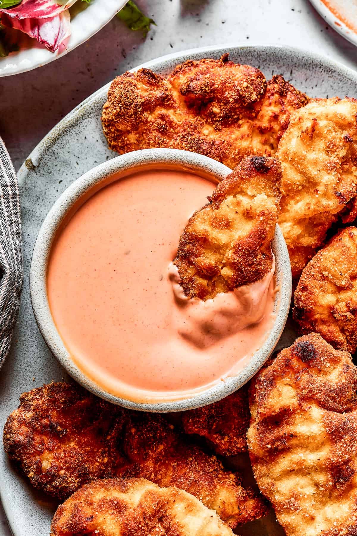 A platter of chicken strips with one piece dipped in a bowl of sauce.