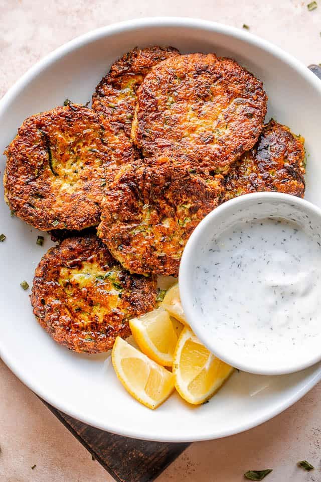 top shot of zucchini cakes in a white bowl with lemon wedges and dip