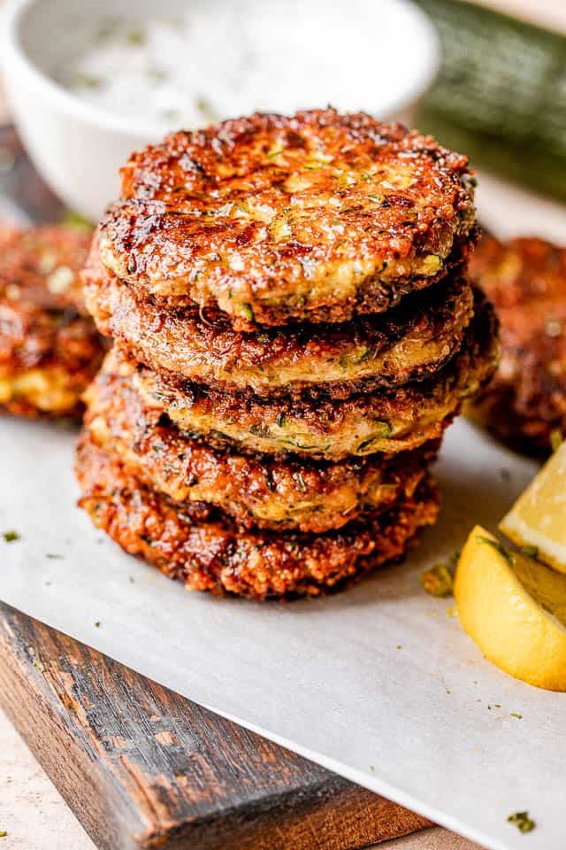 stacked zucchini patties with lemon wedges next to them and yogurt dip behind