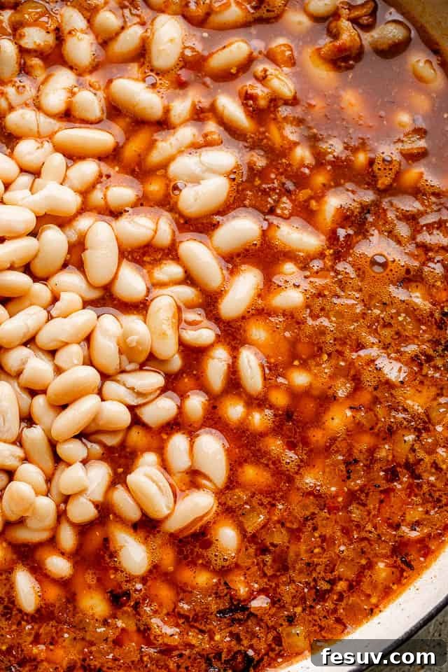 The secret simmers: Beans absorbing root beer, maple, and tangy spices for an unforgettable flavor. Cannellini beans simmering in a rich, dark sauce in a pot, absorbing all the delicious flavors before baking.