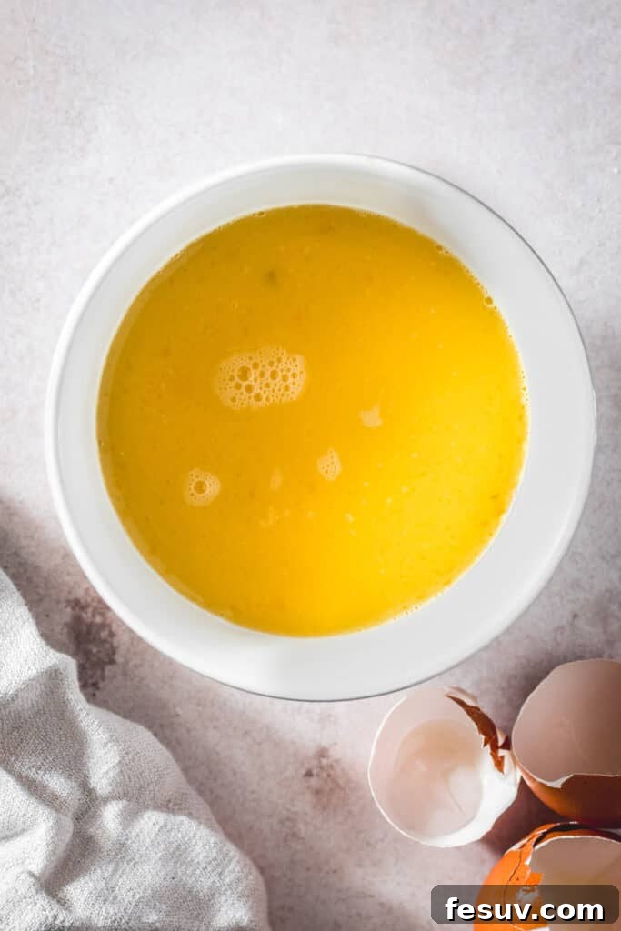 A white mixing bowl with eggs and broth whisked together. Eggshells are nearby on the work surface.
