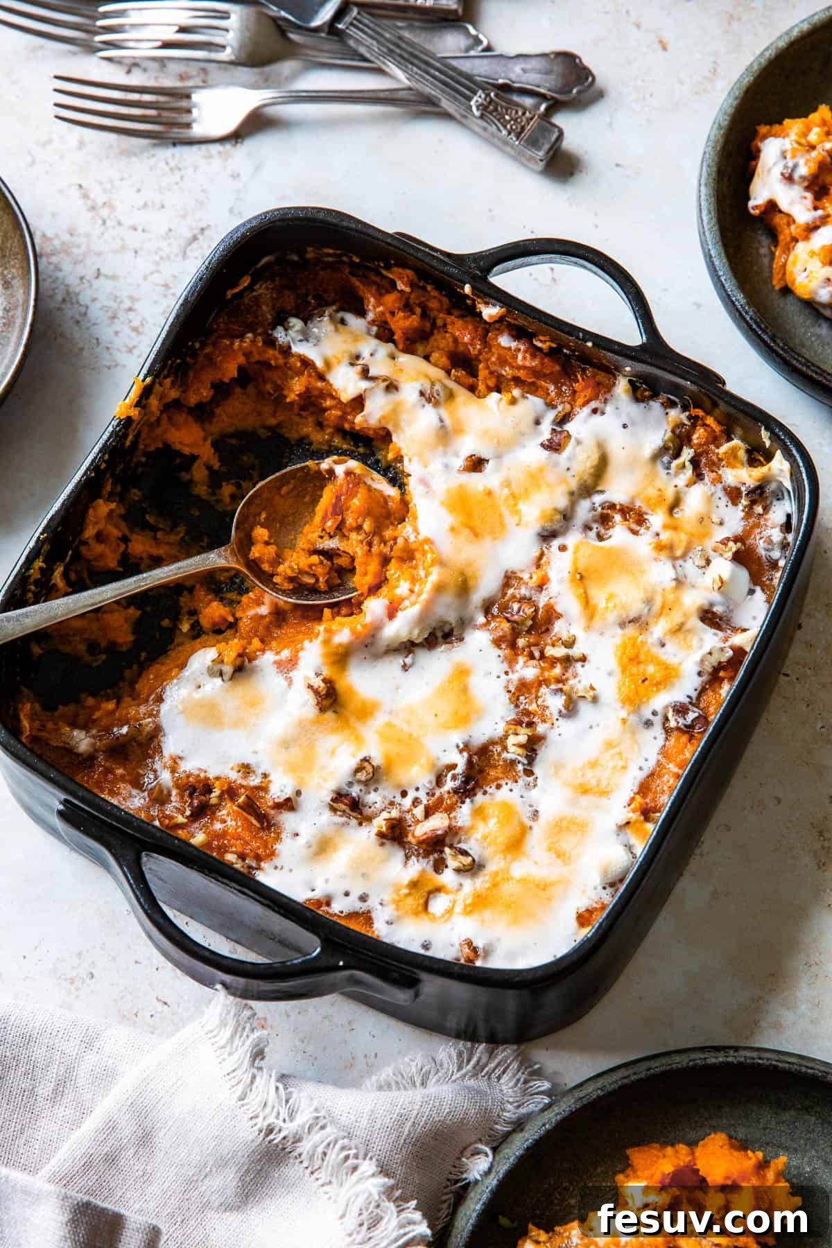 A square black baking dish filled with sweet potato casserole, topped with beautifully melted and golden-brown marshmallows. Several portions have been scooped out, with a serving spoon resting in the dish.