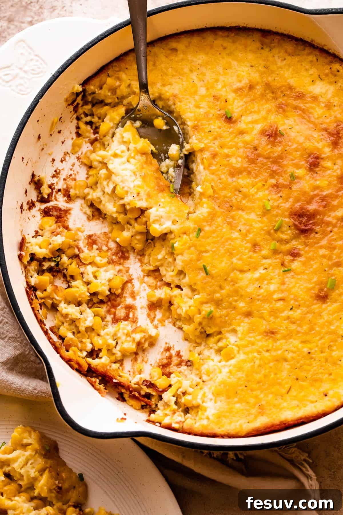 A spoon lifting a serving of golden brown creamed corn pudding from a white cast iron skillet, showcasing its creamy texture.