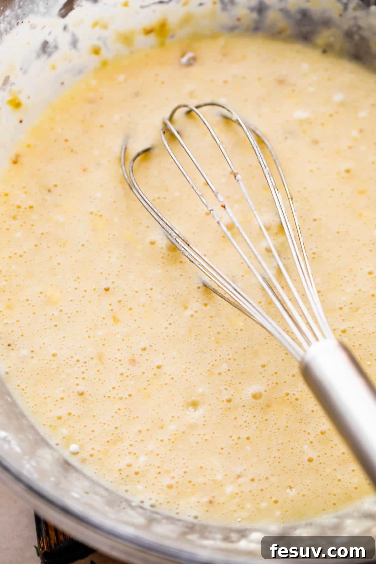Melted butter, sugar, cornstarch, and cream being whisked together in a clear glass mixing bowl, forming the base of the corn pudding batter.