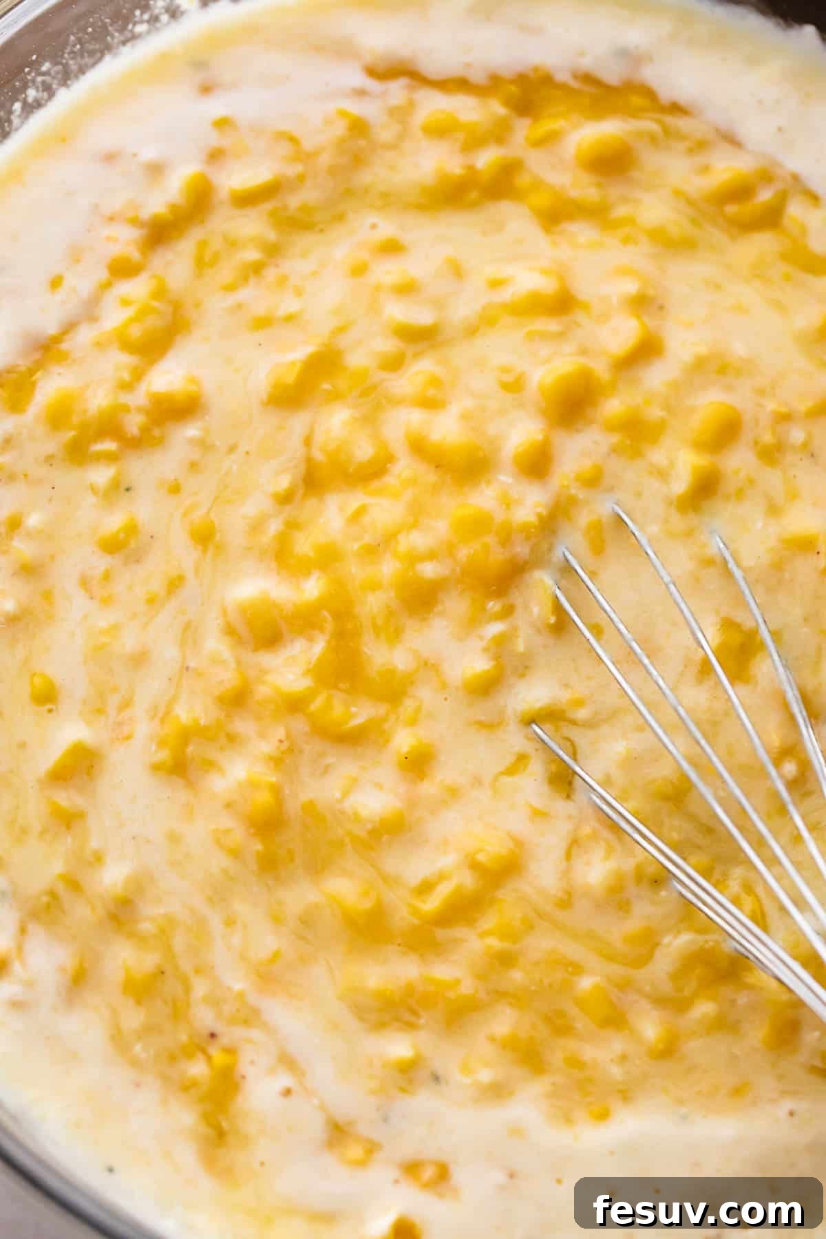 A clear glass mixing bowl filled with smooth, golden corn pudding batter, ready for baking.