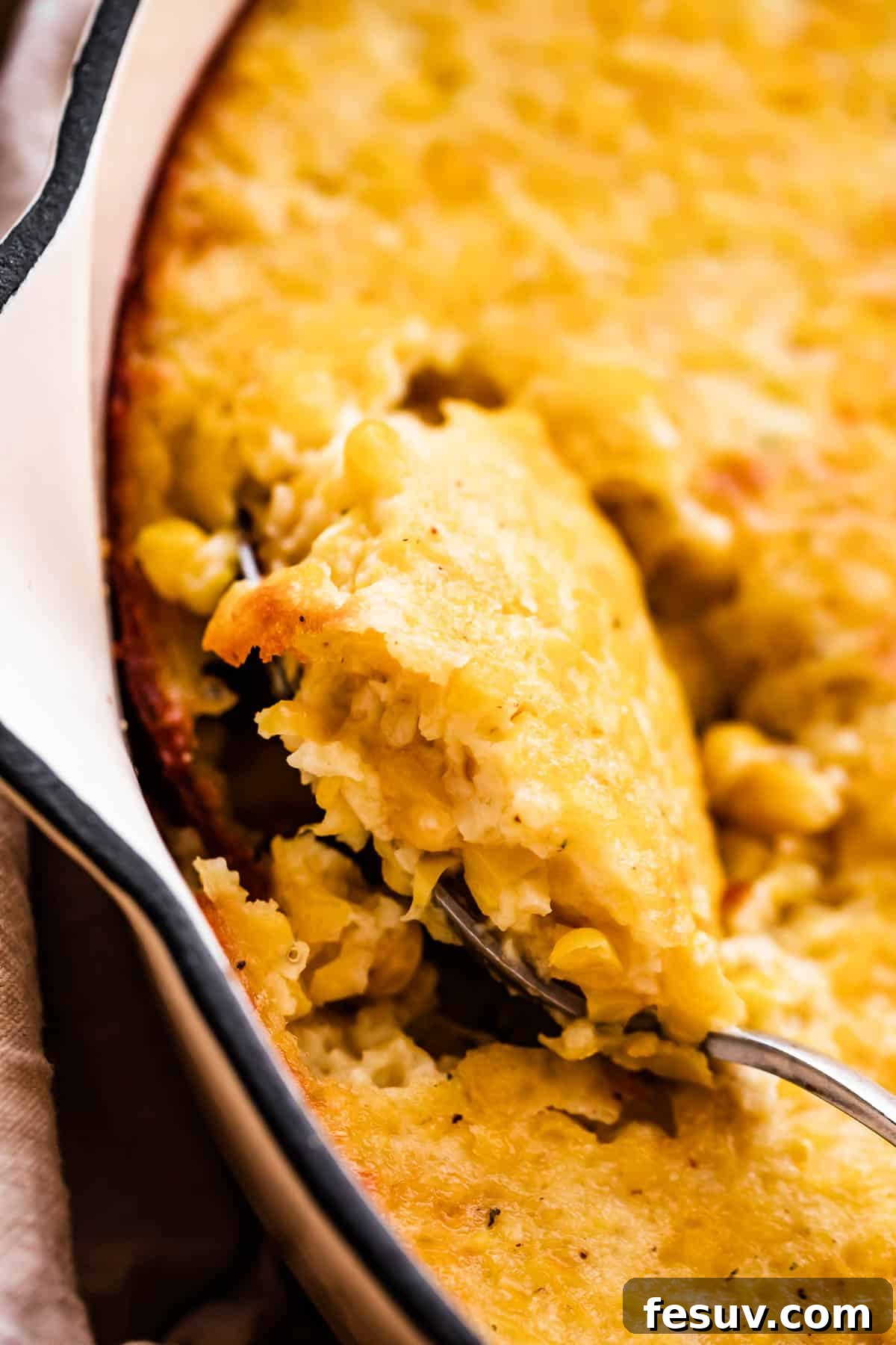 A close-up shot of perfectly baked creamed corn casserole in a white skillet with a serving spoon resting inside.