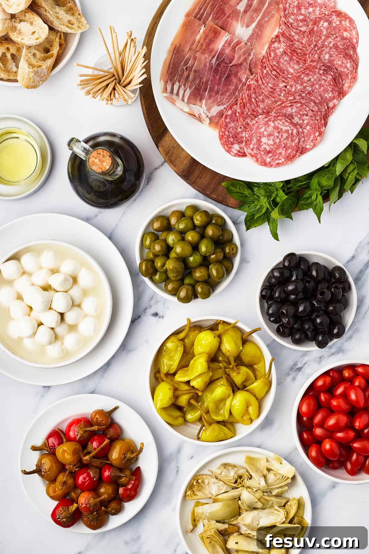 Mini skewers loaded with salami, olives, peppers, and more are stacked and shaped in a ring to make this easy Holiday Antipasto Wreath! A vibrant selection of antipasto ingredients laid out on a table: slices of salami and prosciutto, green and black olives, grape tomatoes, artichoke hearts, pepperoncini, sweet cherry peppers, and fresh mozzarella balls, accompanied by balsamic vinegar, olive oil, and sliced bread.