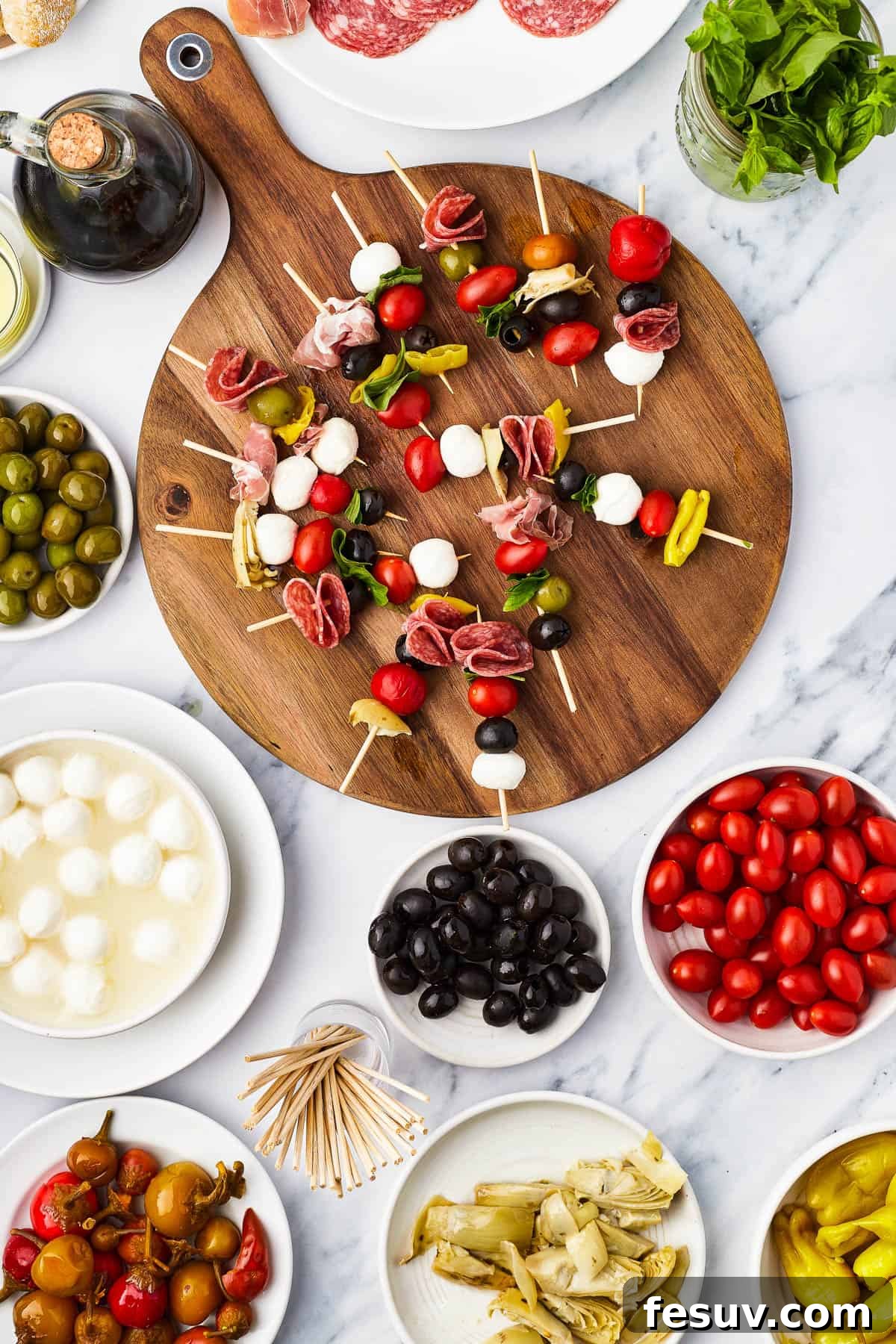 Mini skewers loaded with salami, olives, peppers, and more are stacked and shaped in a ring to make this easy Holiday Antipasto Wreath! An array of prepared antipasto ingredients, including olives, mozzarella, and tomatoes, waiting to be threaded onto skewers on a round cutting board.