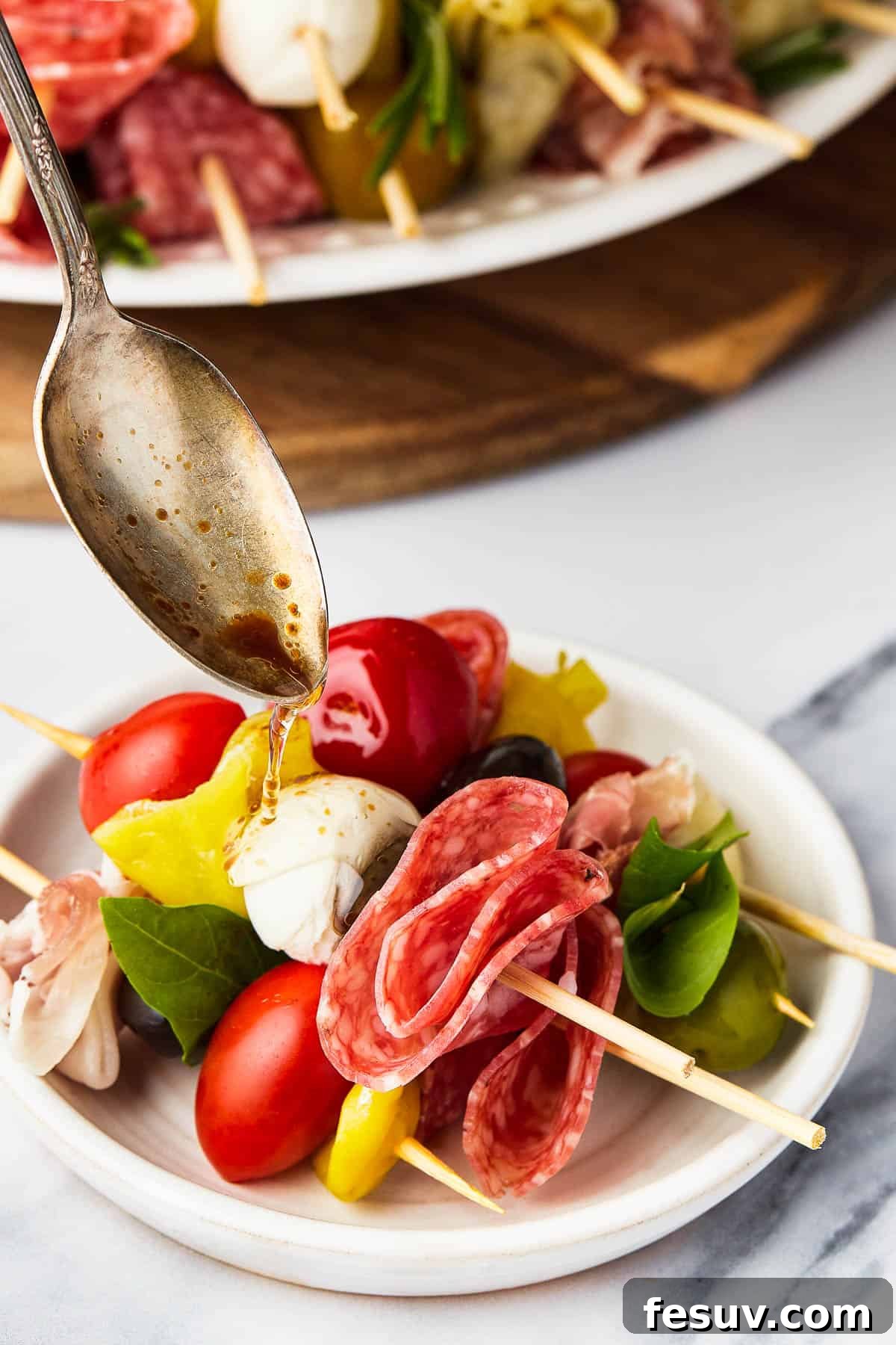 Mini skewers loaded with salami, olives, peppers, and more are stacked and shaped in a ring to make this easy Holiday Antipasto Wreath! A close-up shot of an antipasto skewer being drizzled with balsamic vinegar, highlighting the fresh ingredients.