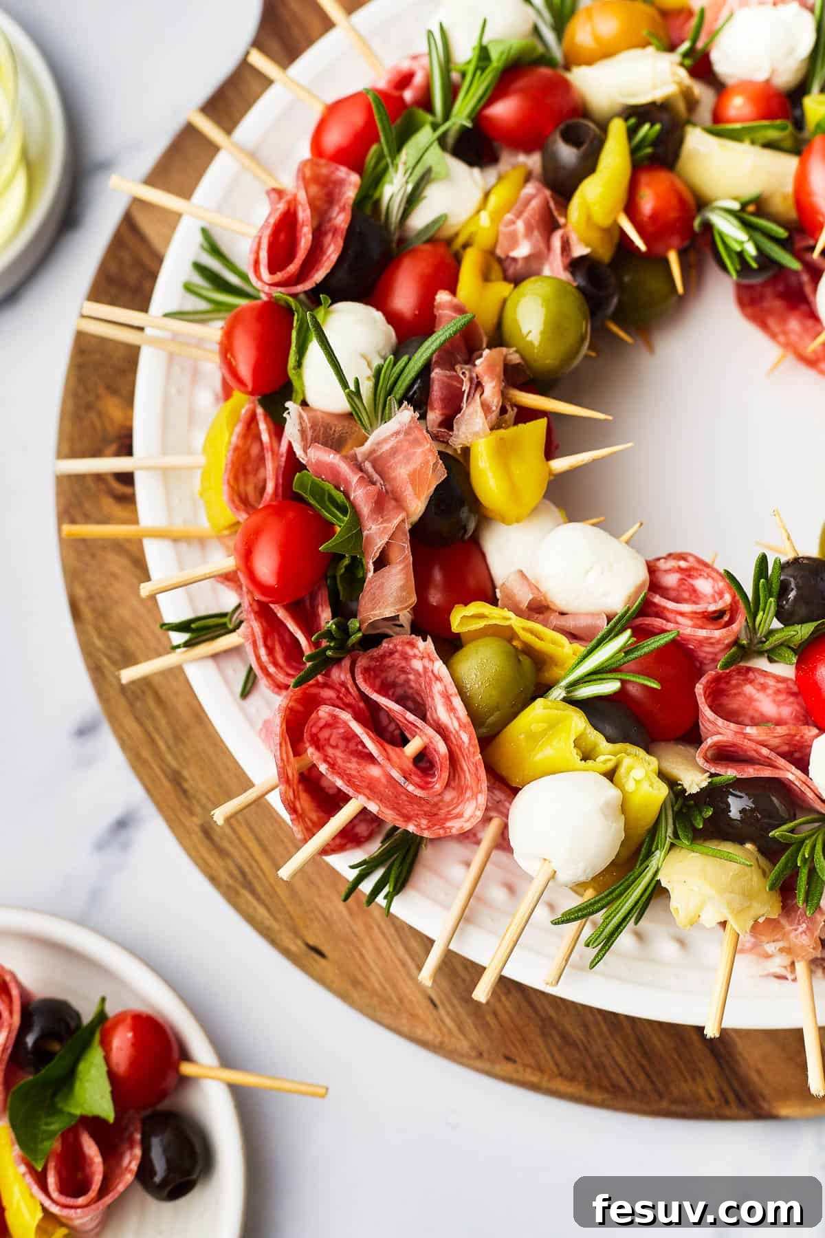 Mini skewers loaded with salami, olives, peppers, and more are stacked and shaped in a ring to make this easy Holiday Antipasto Wreath! A close-up view of the Holiday Antipasto Wreath, showcasing the vibrant skewers of prosciutto, pepperoncini, grape tomatoes, mozzarella, and olives, garnished with fresh rosemary.