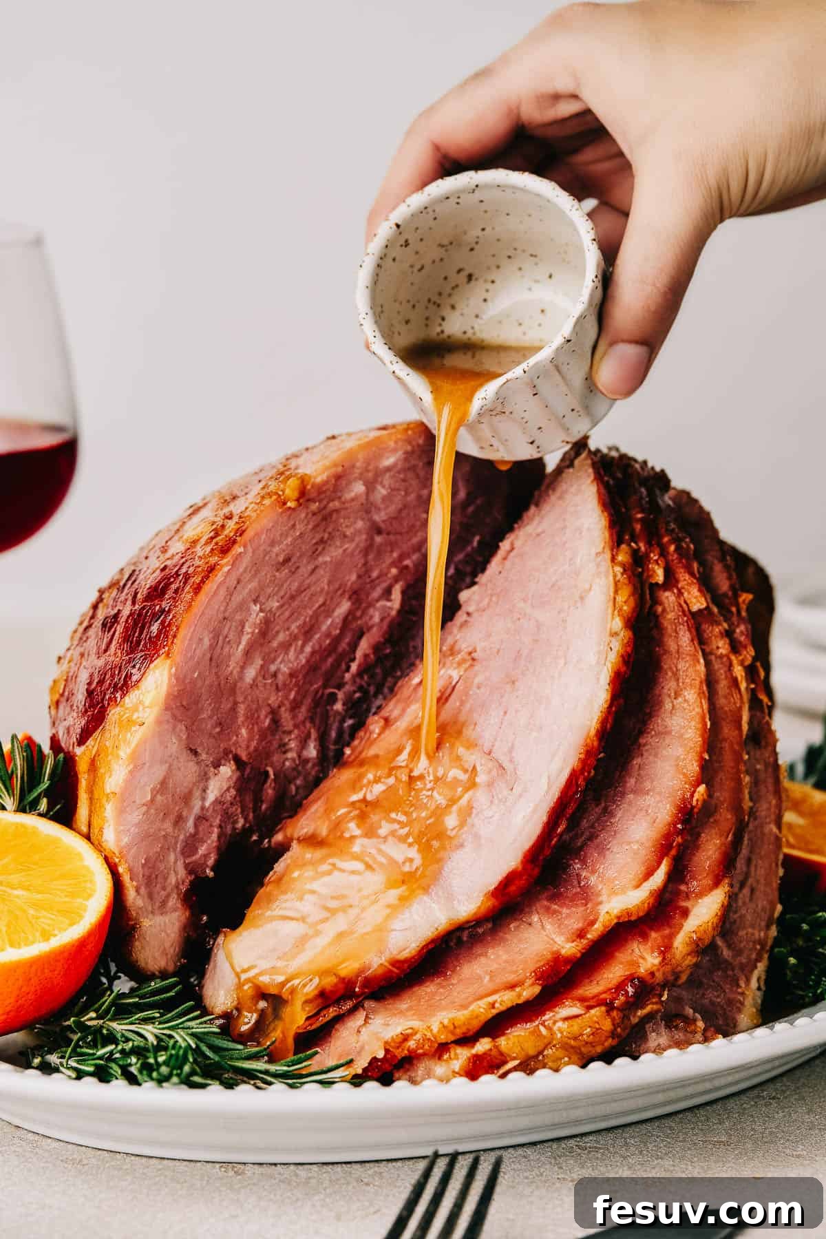 A woman's hand is pouring glaze from a small white measuring cup over a cooked spiral ham on a serving platter.