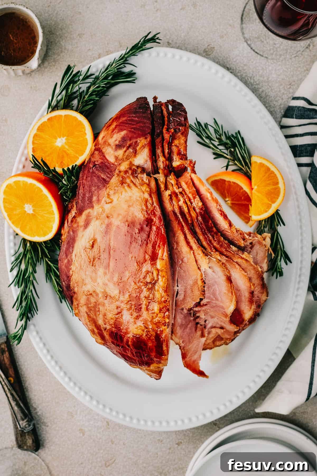 Overhead shot of a partially carved ham, with slices fanning out, on a serving platter garnished with orange slices and rosemary.