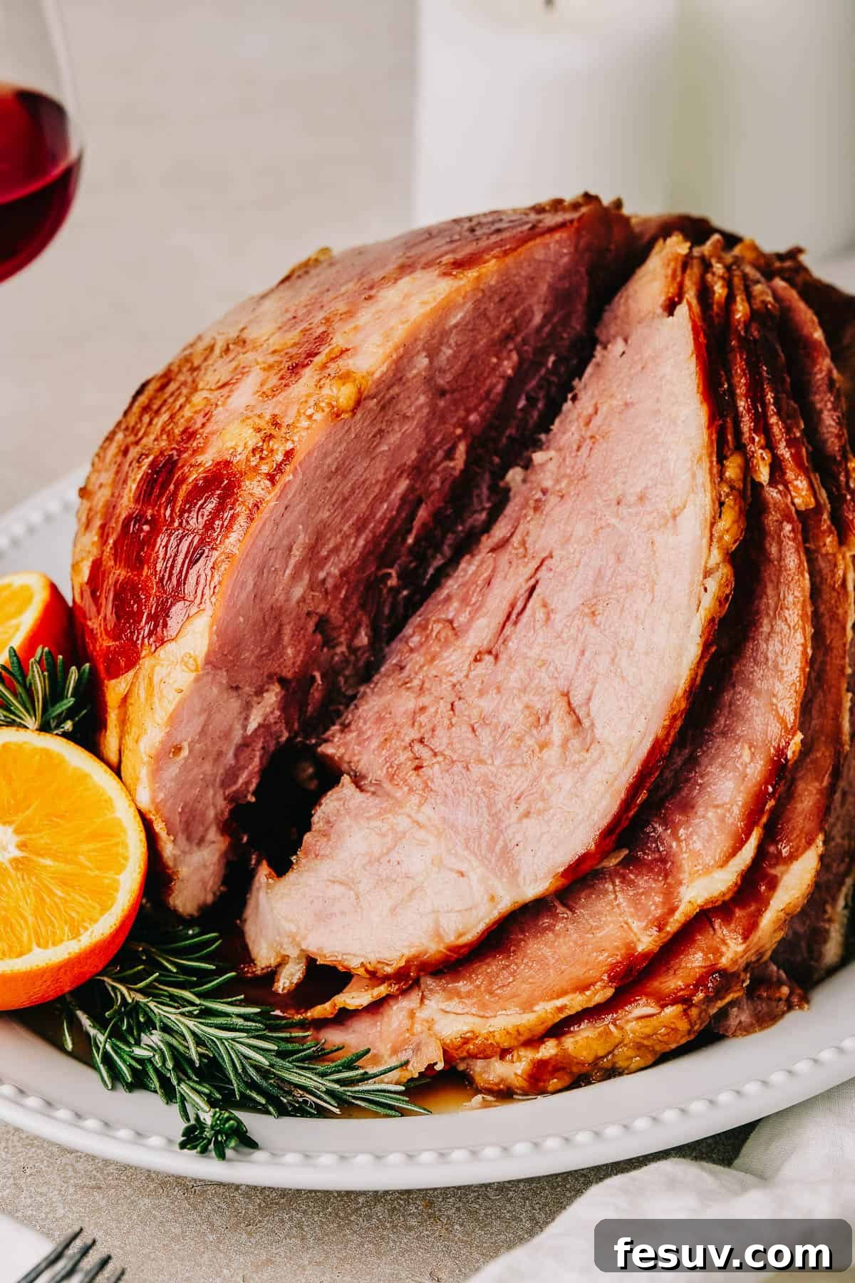 Sliced ham on a serving platter. A candle and a glass of wine are in the background of the shot, while a cup of glaze and several forks are in the foreground.