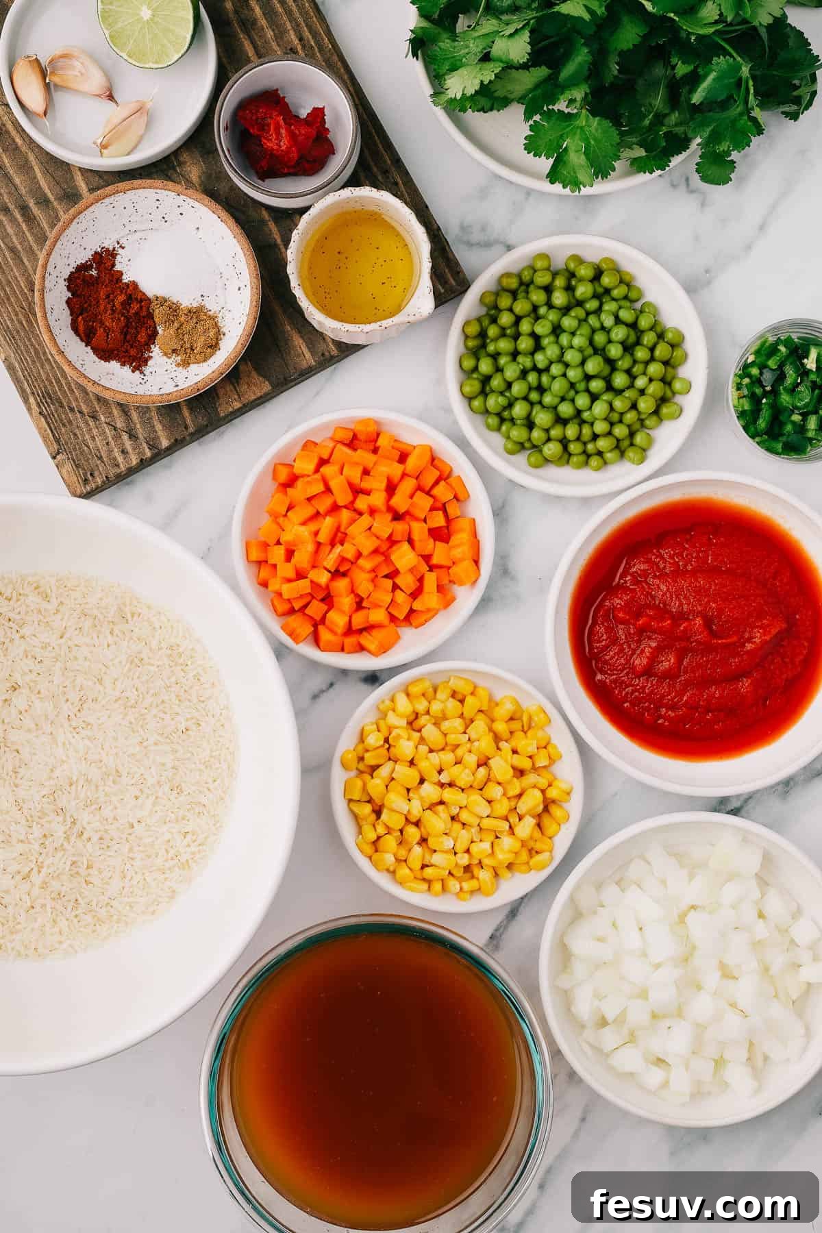 From left to right, a selection of fresh and pantry ingredients for Instant Pot Mexican Rice, including garlic, lime, tomato paste, cilantro, chili powder, salt, cumin, avocado oil, green peas, a whole jalapeno, uncooked white rice, diced carrots, corn kernels, tomato puree, vegetable broth, and a white onion.