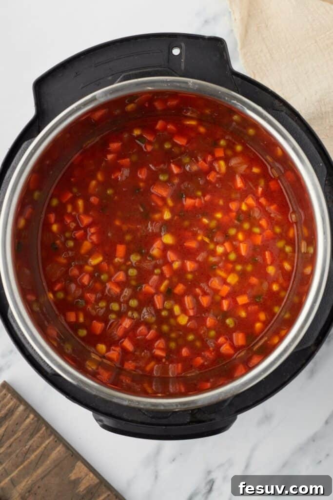 Overhead shot of an Instant Pot, with all raw ingredients, including rinsed rice, vegetables, and tomato-based broth, stirred together before pressure cooking.